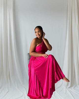 Woman wearing one-shoulder blush pink pleated maxi dress with flowing skirt — elegant evening gown from Tulley’s Boutique.