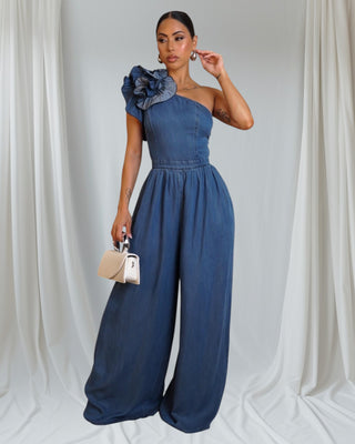 Woman wearing a blue one-shoulder jumpsuit with a floral detail against a white curtain background
