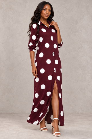 Model touching the pointed collar of a burgundy and white polka dot maxi shirt dress with a front slit