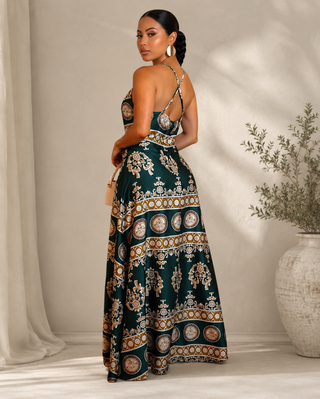 A woman wearing a dark green halter-neck maxi dress with an intricate gold and beige bohemian-style floral and geometric pattern, standing in a sunlit room with a neutral beige background.