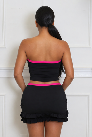 A woman shown from the back wearing a black two-piece set consisting of a strapless bandeau crop top with a bright pink band and matching high-waisted ruffled mini skirt.