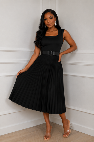 A woman modeling a sleeveless black midi dress with a square neckline, a thin black belt at the waist, and a pleated skirt, standing against a white paneled wall.