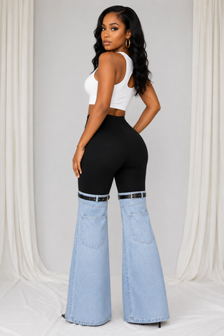 A woman modeling a pair of high-waisted flared jeans with a black upper section and light blue denim lower legs, styled with a white crop top against a draped white fabric backdrop.