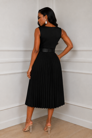 A woman modeling a sleeveless black midi dress with a pleated skirt and a thin black belt at the waist, shown from the back with pockets