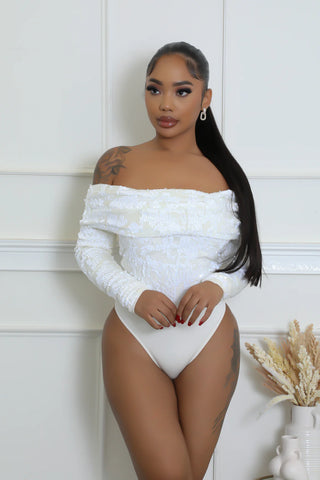 Woman wearing a white off-shoulder bodysuit against a white wall.
