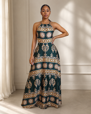 A woman modeling a sleeveless halter-neck maxi dress with an intricate gold, beige, and cream circular medallion pattern on a dark teal-green background, standing in a brightly lit room with warm sunlight and shadows.