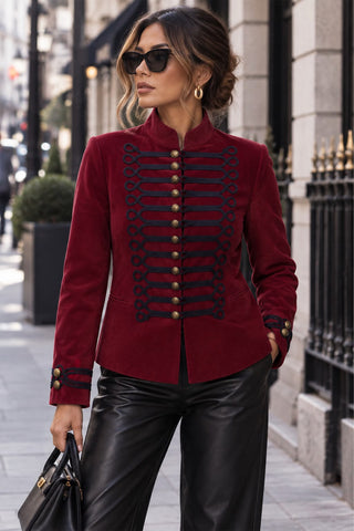 Model wearing a red velvet military band jacket with black cord frog trim, gold buttons and stand collar, styled with black leather pants
