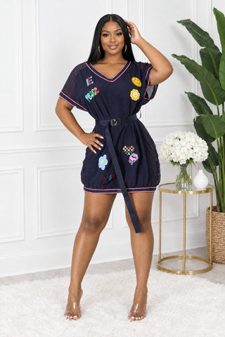 Model wearing a navy jersey mini dress with retro patches, pink striped trim and a buckle belt