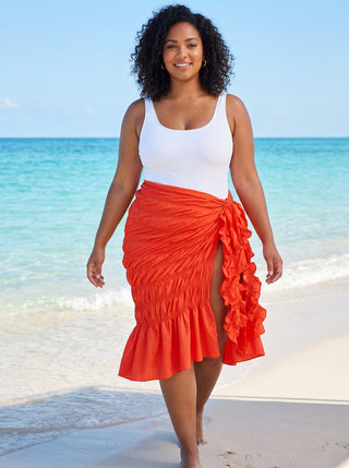 Model walking barefoot at the water's edge in an orange ruched ruffle wrap skirt cover-up with front slit