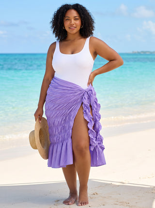 Lavender purple ruffle slit beach wrap skirt with self-tie hip knot worn over a white swimsuit, model holding a straw sun hat