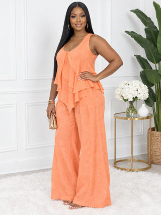 Three-quarter view of model with hand on hip in the orange linen two-piece set, showing the draped asymmetric layers of the top and the flowing wide-leg pants.
