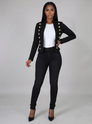Model wearing a black cropped military band jacket with gold domed buttons and black cord trim, open over a white top and black skinny jeans