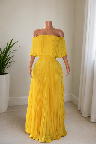 A bright yellow off-the-shoulder maxi dress with a pleated design, displayed on a mannequin in an indoor setting with a potted plant and curtains.